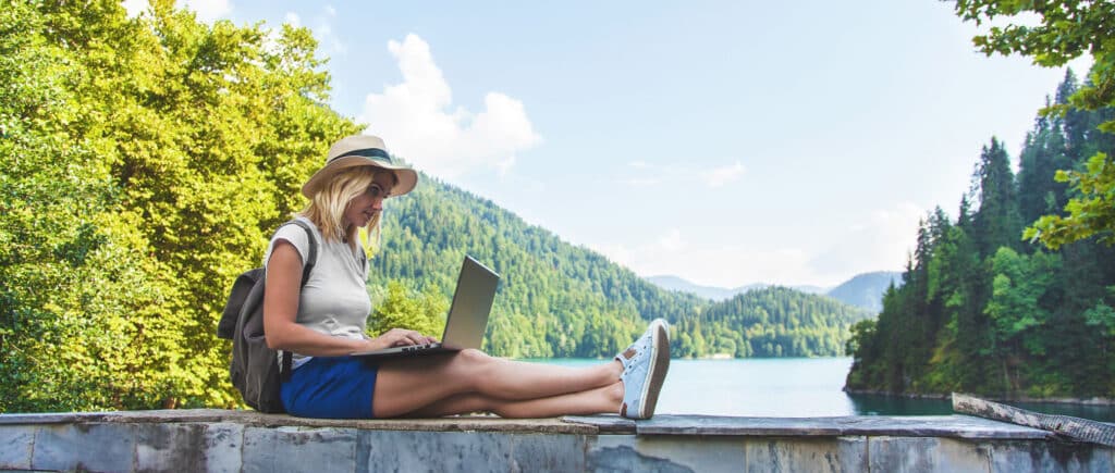 Beautiful girl traveler in a hat on a lake with a laptop working on a ledge